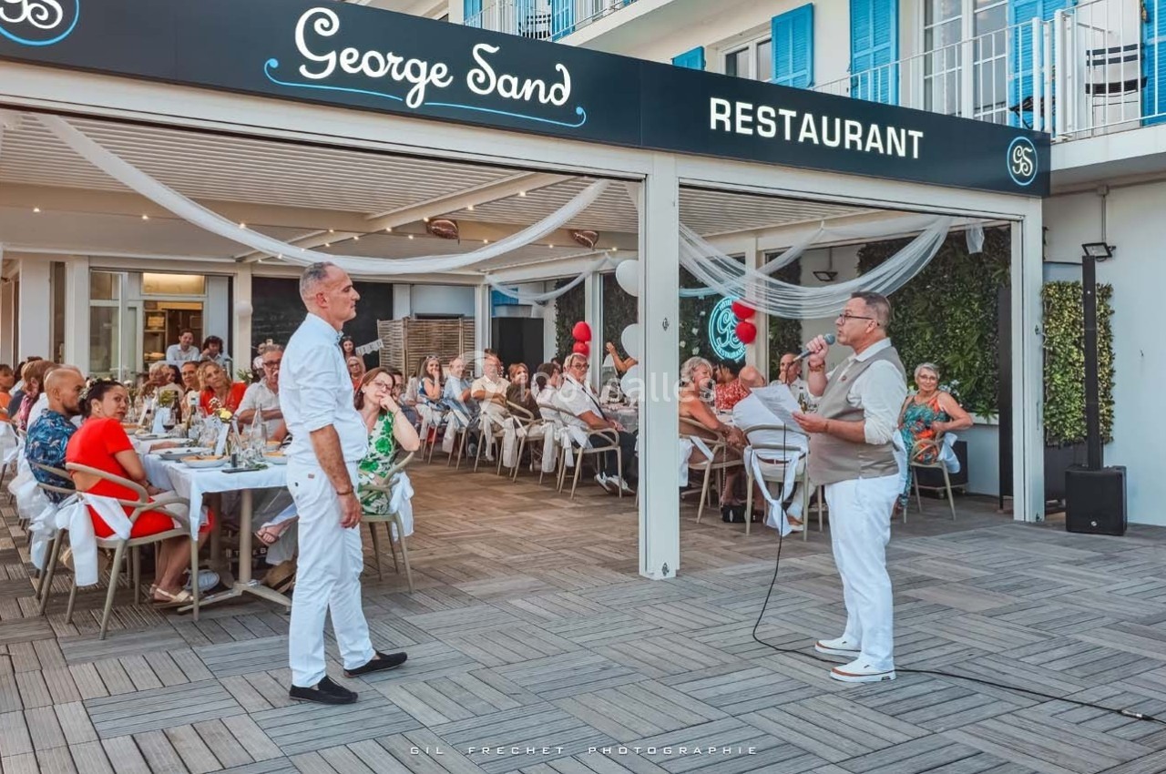 Un homme s'adresse à un groupe de personnes assises à des tables en terrasse devant un restaurant nommé ’George Sand’.