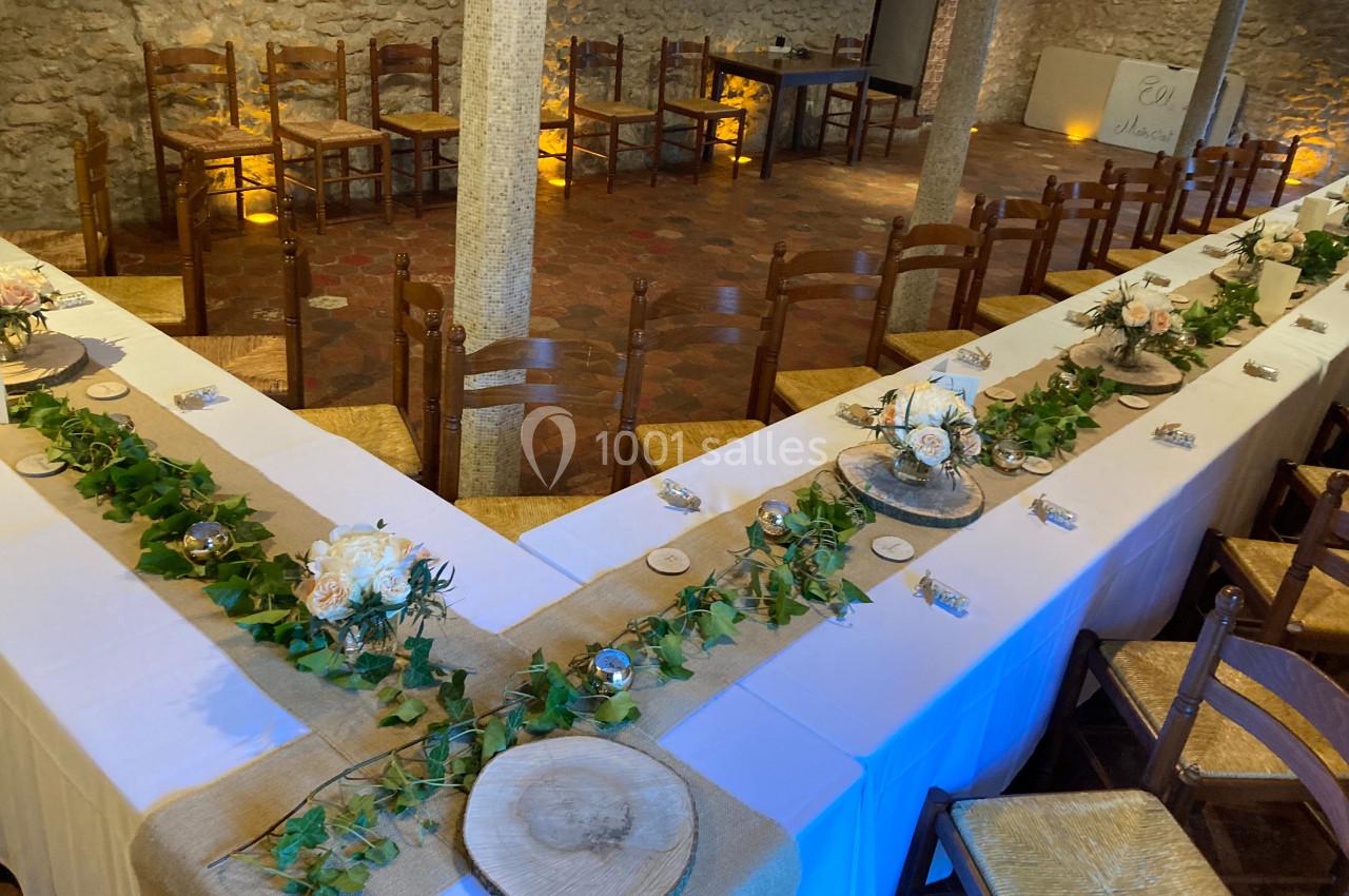 Tables en U décorées de nappes blanches, chemins de table en toile de jute, feuillage et fleurs, dans une salle en pierre.