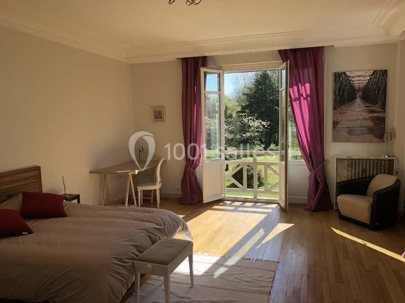 Chambre lumineuse avec lit double, bureau, fauteuil, rideaux violets et porte-fenêtre donnant sur un jardin verdoyant.
