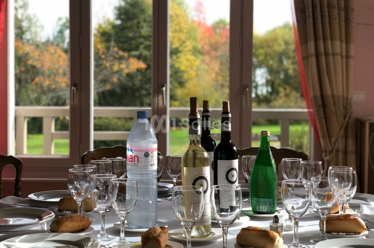 Table dressée avec des verres, bouteilles de vin, eau plate et gazeuse, et petits pains, devant une fenêtre donnant sur un…