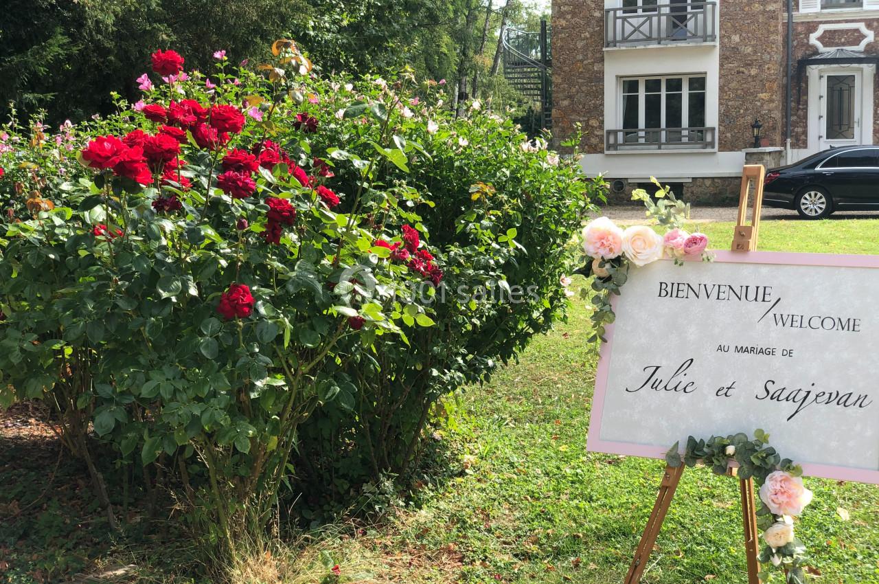 Panneau de bienvenue à un mariage, posé sur un chevalet près d'un massif de roses dans un jardin devant une maison.