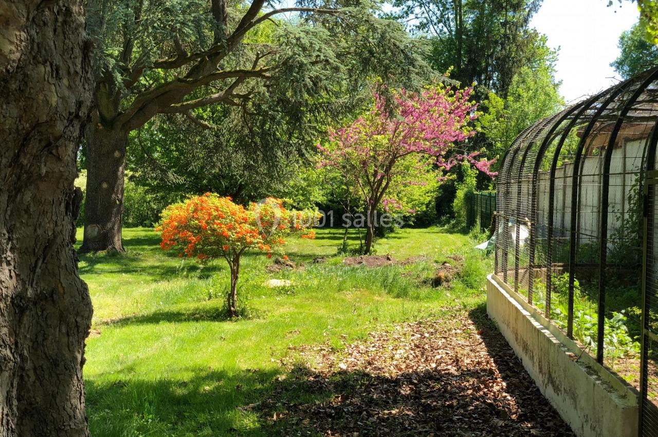 Jardin verdoyant avec arbres fleuris, arbuste orange et serre en verre à droite, éclairé par une lumière naturelle.