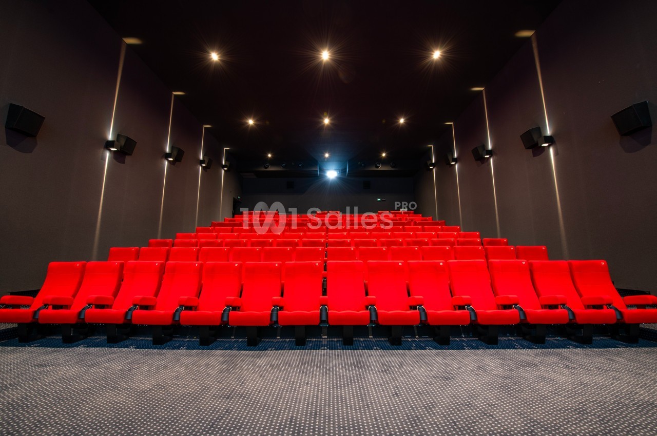 Salle de cinéma vide avec des rangées de fauteuils rouges alignés face à un écran blanc.