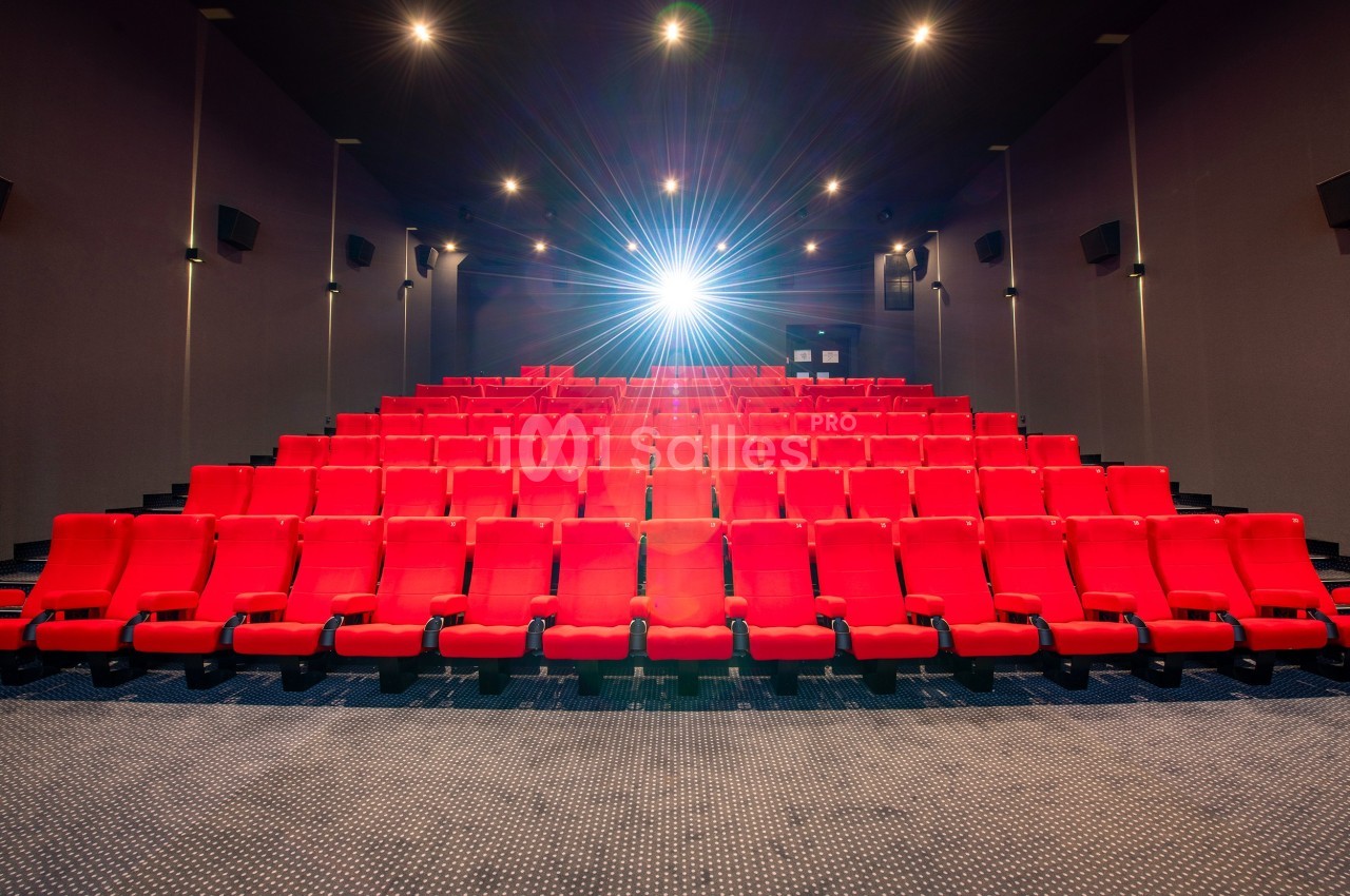 Salle de cinéma vide avec des rangées de fauteuils rouges et un projecteur allumé au fond.