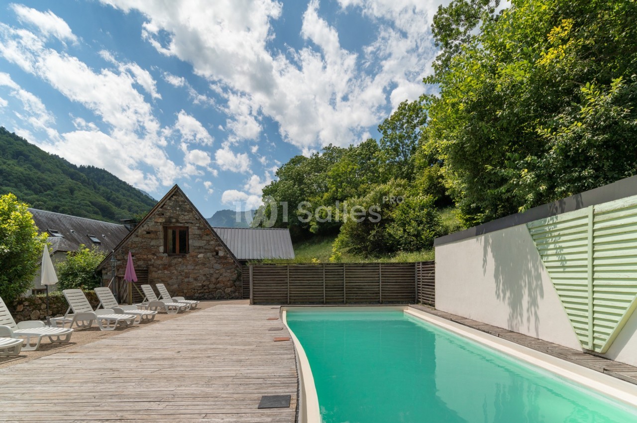 Piscine extérieure bordée de transats sur une terrasse en bois, entourée de verdure et de montagnes.