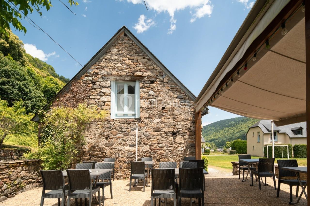 Terrasse extérieure avec tables et chaises devant une maison en pierre, entourée de verdure et sous un ciel dégagé.
