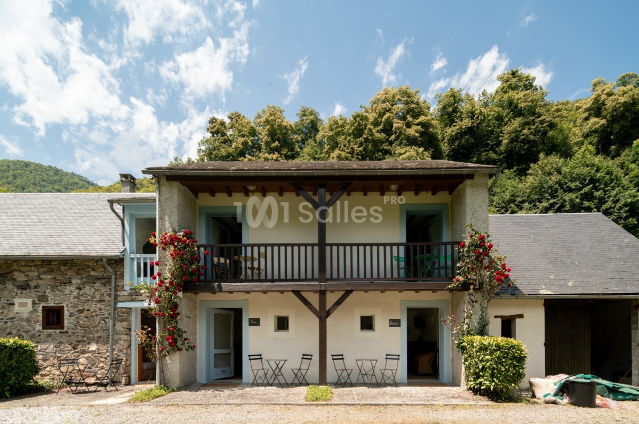 Façade d'une maison rurale à deux étages avec balcon en bois, entourée de végétation et de rosiers grimpants.