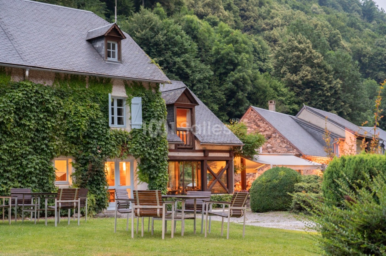 Façade d'une maison en pierre couverte de lierre, entourée de verdure avec des tables et chaises en extérieur.