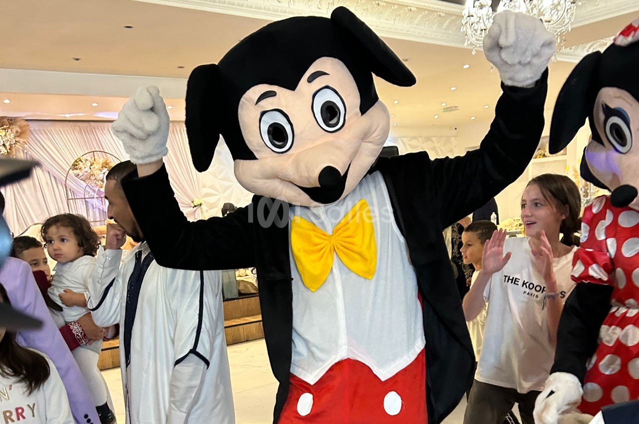 Personne déguisée en souris anthropomorphe avec un nœud jaune et un costume rouge, entourée d'enfants dans un lieu festif.