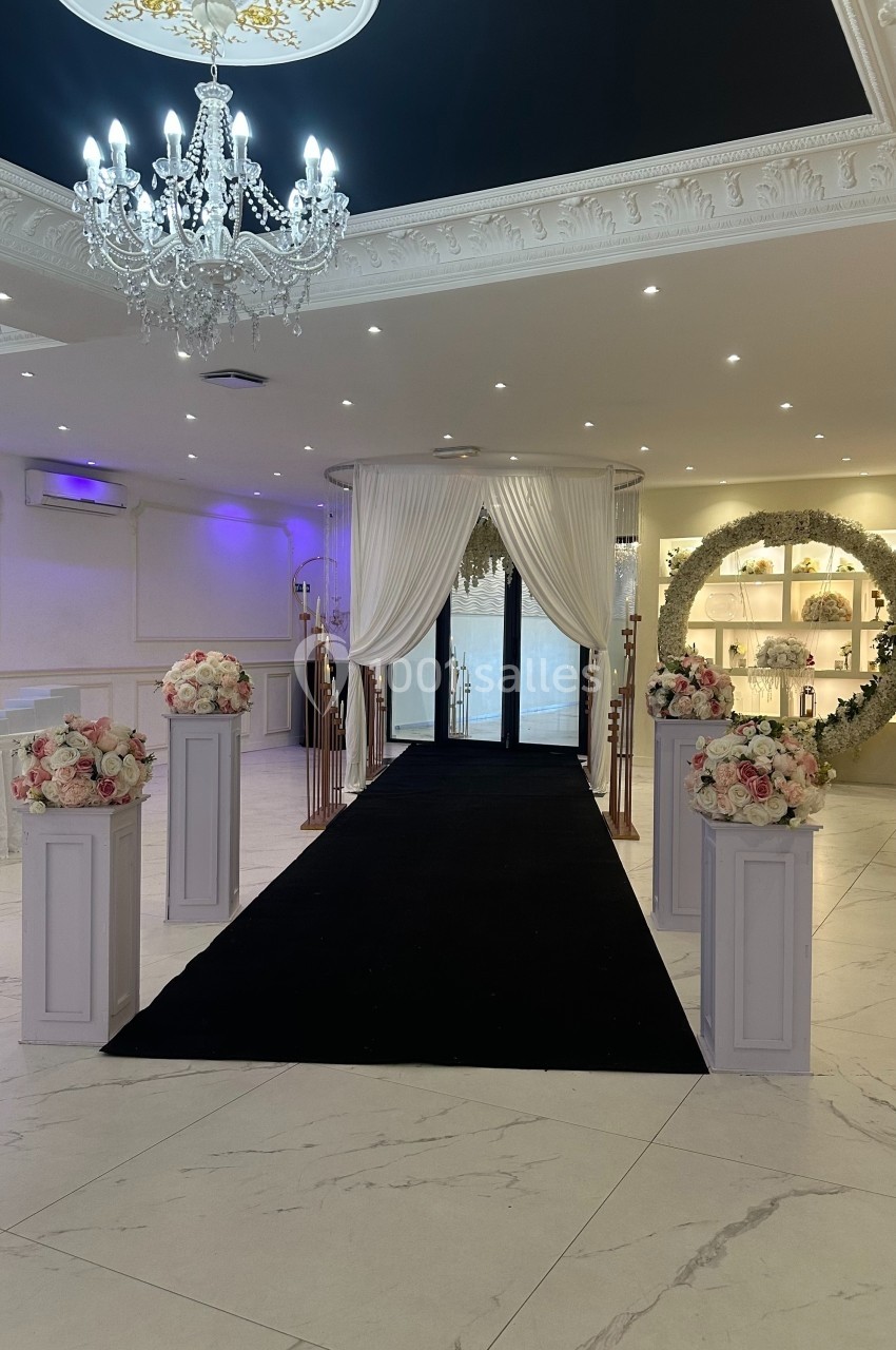 Allée décorée avec tapis noir, colonnes fleuries, rideaux blancs et arche florale dans une salle élégante.