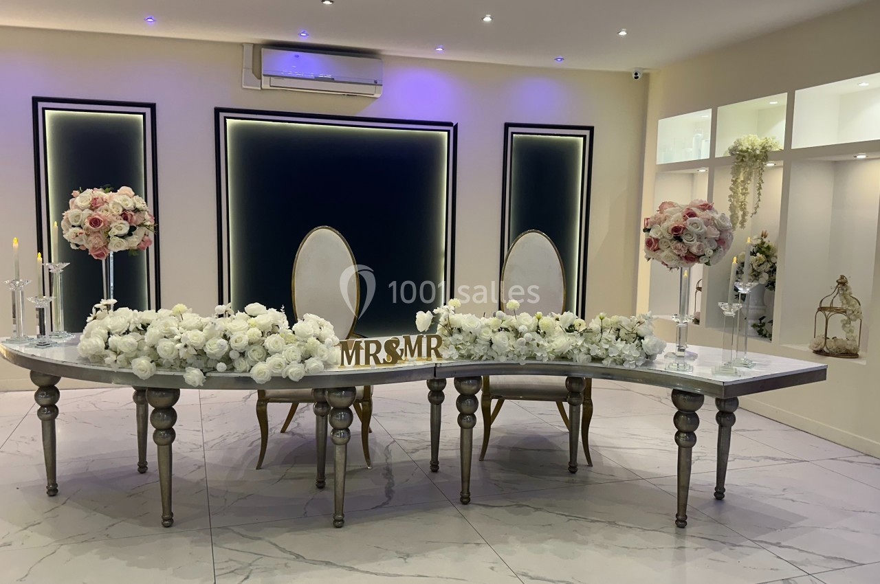 Table de mariage décorée de fleurs blanches et roses, avec deux chaises et un panneau ’Mr & Mrs’ au centre.