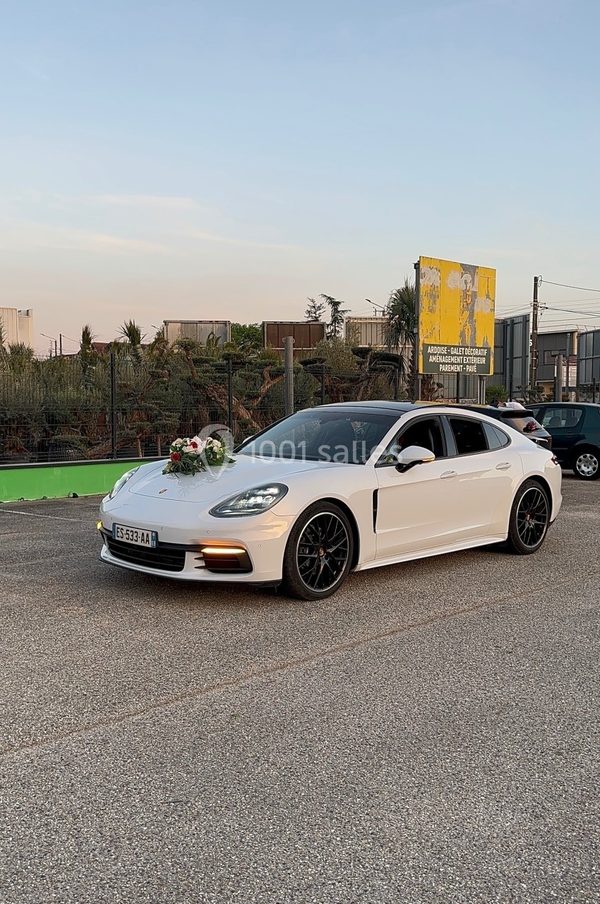 Voiture blanche décorée de fleurs à l'avant, stationnée sur une route près d'une clôture et d'un panneau publicitaire.