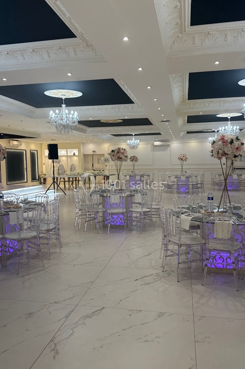 Salle de réception élégante avec tables décorées de fleurs, chaises transparentes et éclairage violet au sol.