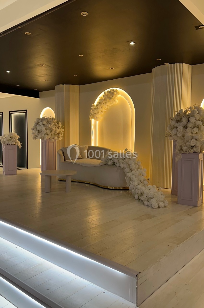 Scène décorée avec un canapé beige, colonnes et arrangements floraux blancs sous un éclairage chaleureux.