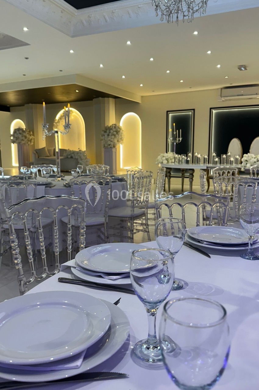 Salle de réception élégante avec tables dressées, chaises transparentes et décoration lumineuse.