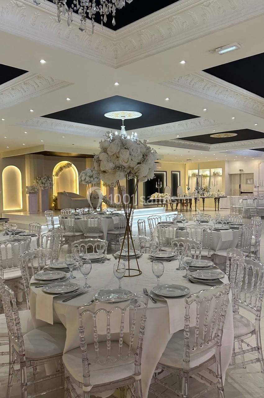 Salle de réception élégante avec tables rondes dressées, chaises transparentes et décoration florale blanche.