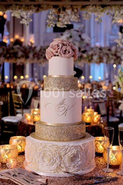 Gâteau de mariage à quatre étages décoré de paillettes dorées, fleurs roses et bougies en arrière-plan.