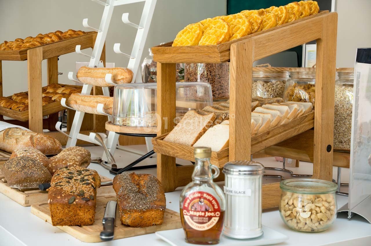 Buffet de petit-déjeuner avec pains variés, céréales, gaufres, noix et condiments disposés sur des étagères en bois.
