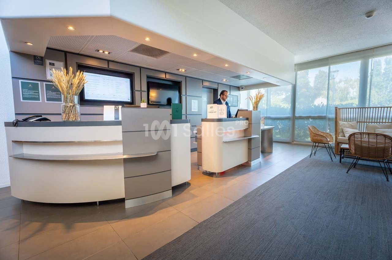 Accueil d'un hôtel avec un comptoir moderne, un employé à l'arrière-plan et un espace d'attente lumineux.