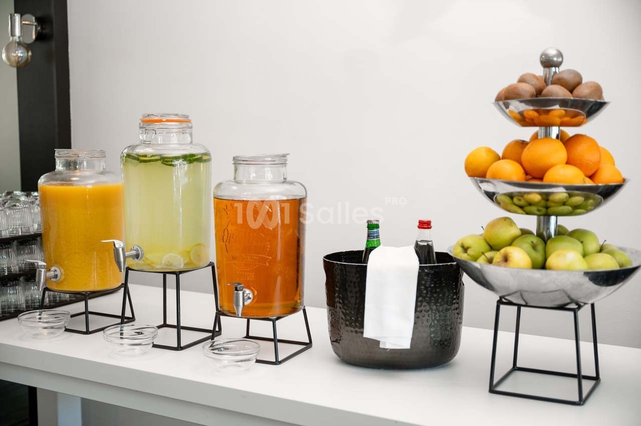 Distributeurs de jus et d'eau citronnée, bol avec boissons fraîches et corbeilles de fruits sur une table blanche.