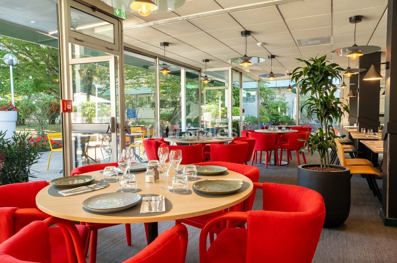 Salle de restaurant lumineuse avec des tables dressées, des chaises rouges et une vue sur une terrasse verdoyante.