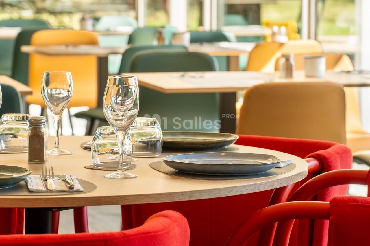 Salle de restaurant lumineuse avec tables dressées, chaises colorées et vue sur l'extérieur verdoyant.