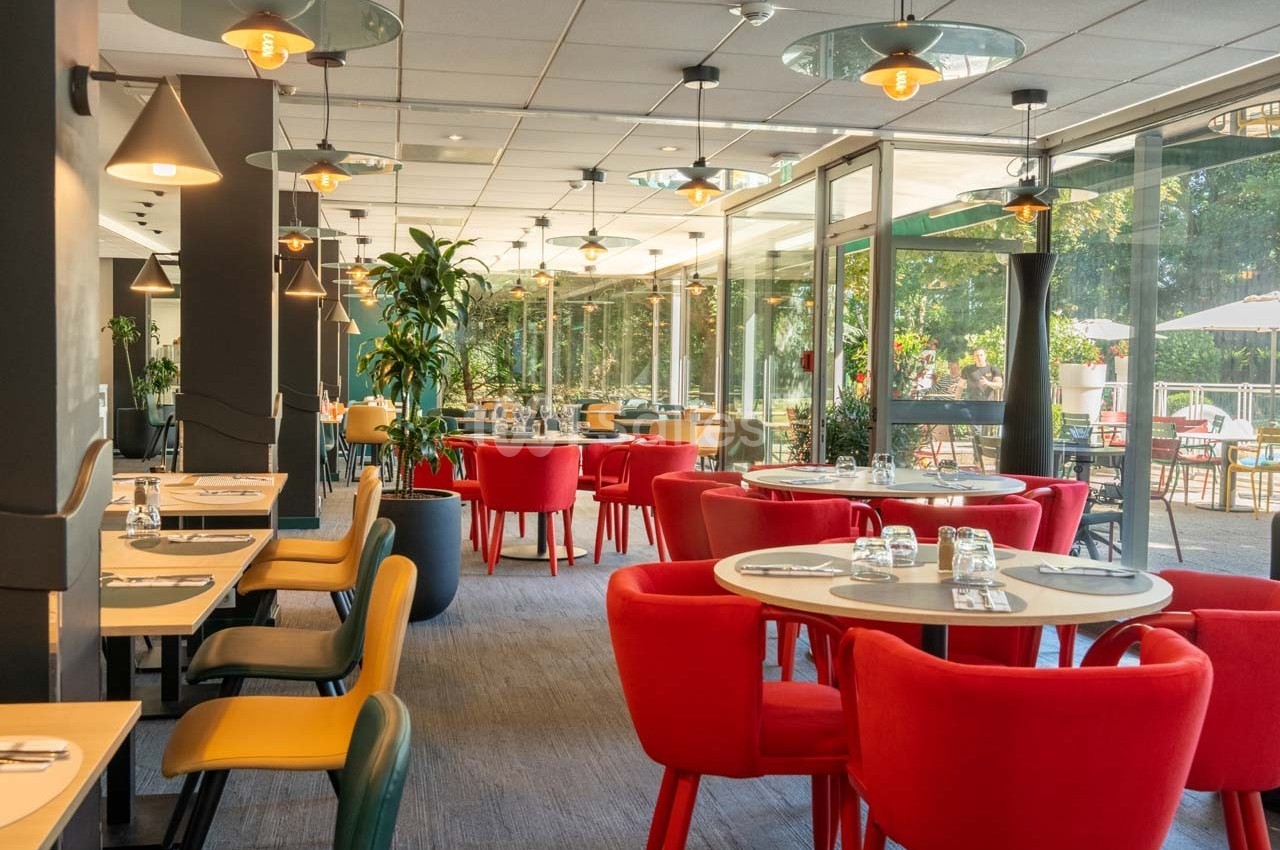 Salle de restaurant lumineuse avec des tables dressées, des chaises colorées et de grandes baies vitrées donnant sur un…
