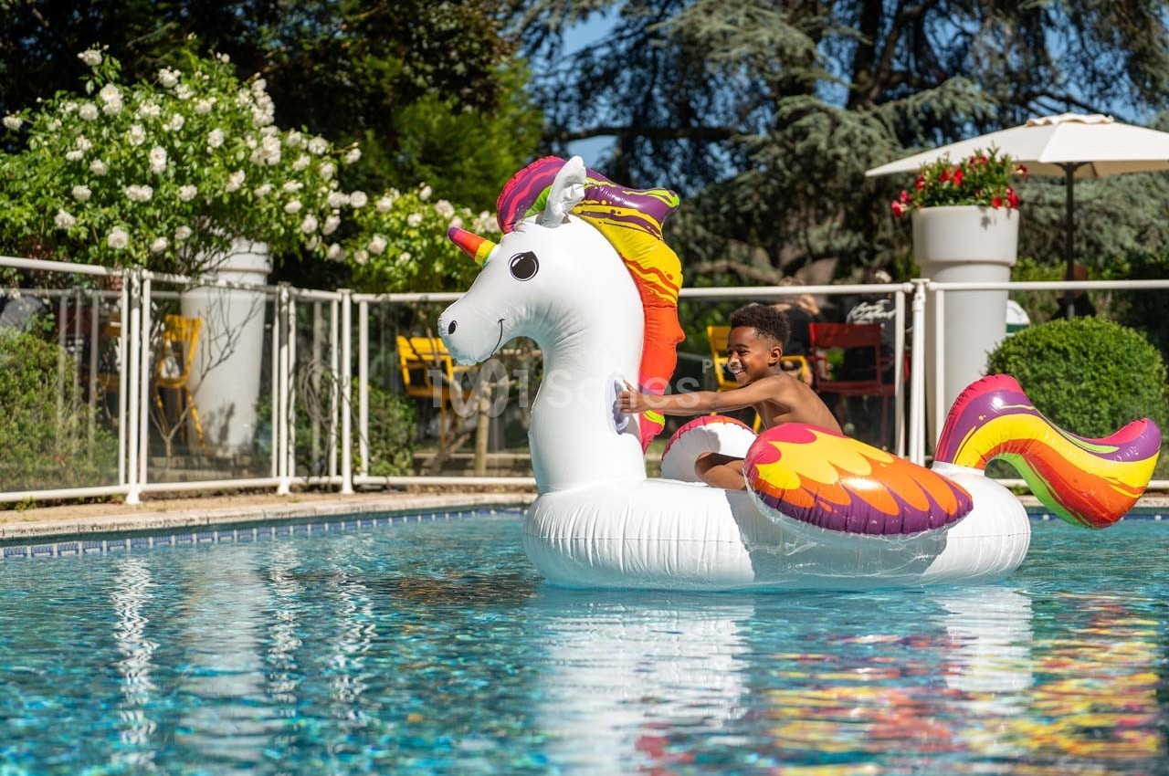 Un enfant souriant joue sur une bouée licorne colorée dans une piscine extérieure entourée de végétation.