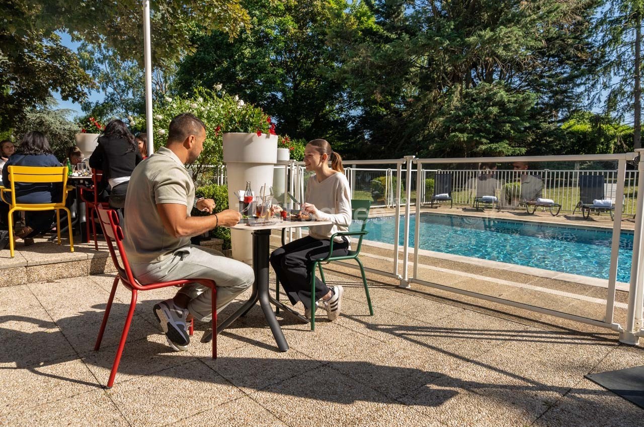Deux personnes dînent en terrasse près d'une piscine, entourées de chaises colorées et de végétation.