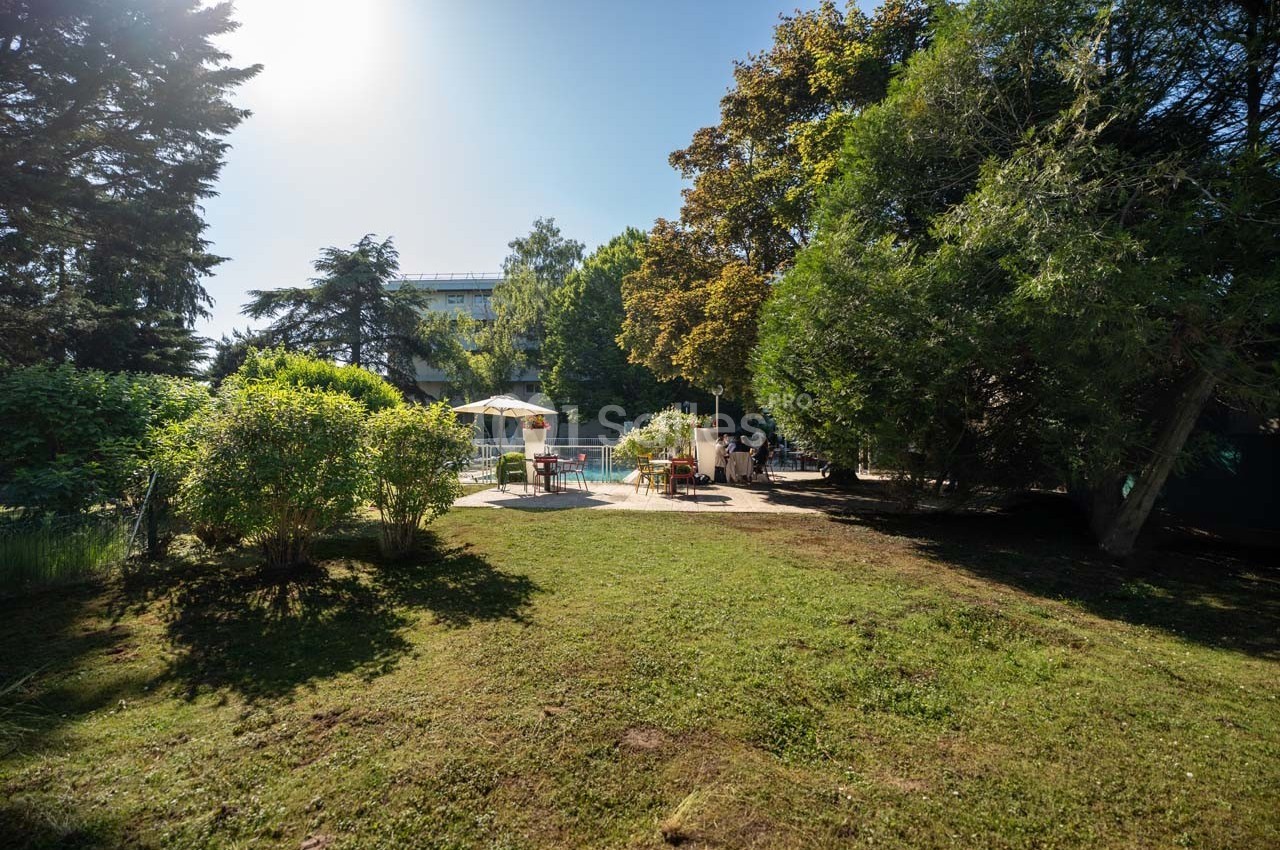 Espace extérieur verdoyant avec pelouse, arbres et une piscine entourée de chaises longues sous un ciel dégagé.