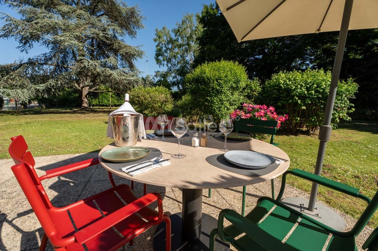 Table de jardin avec chaises colorées, couverts dressés, sous un parasol, entourée de verdure et fleurs en arrière-plan.