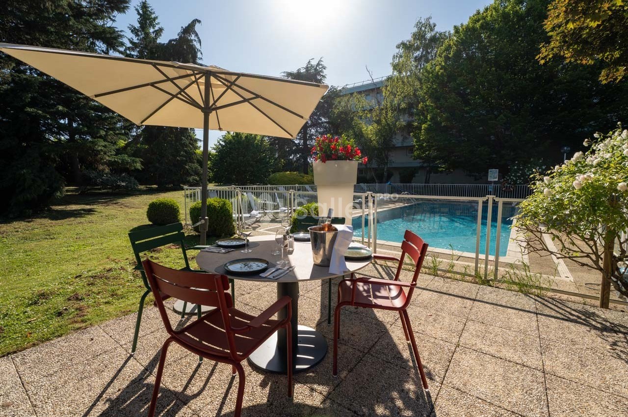 Table dressée en terrasse avec parasol, vue sur une piscine entourée de verdure par une journée ensoleillée.