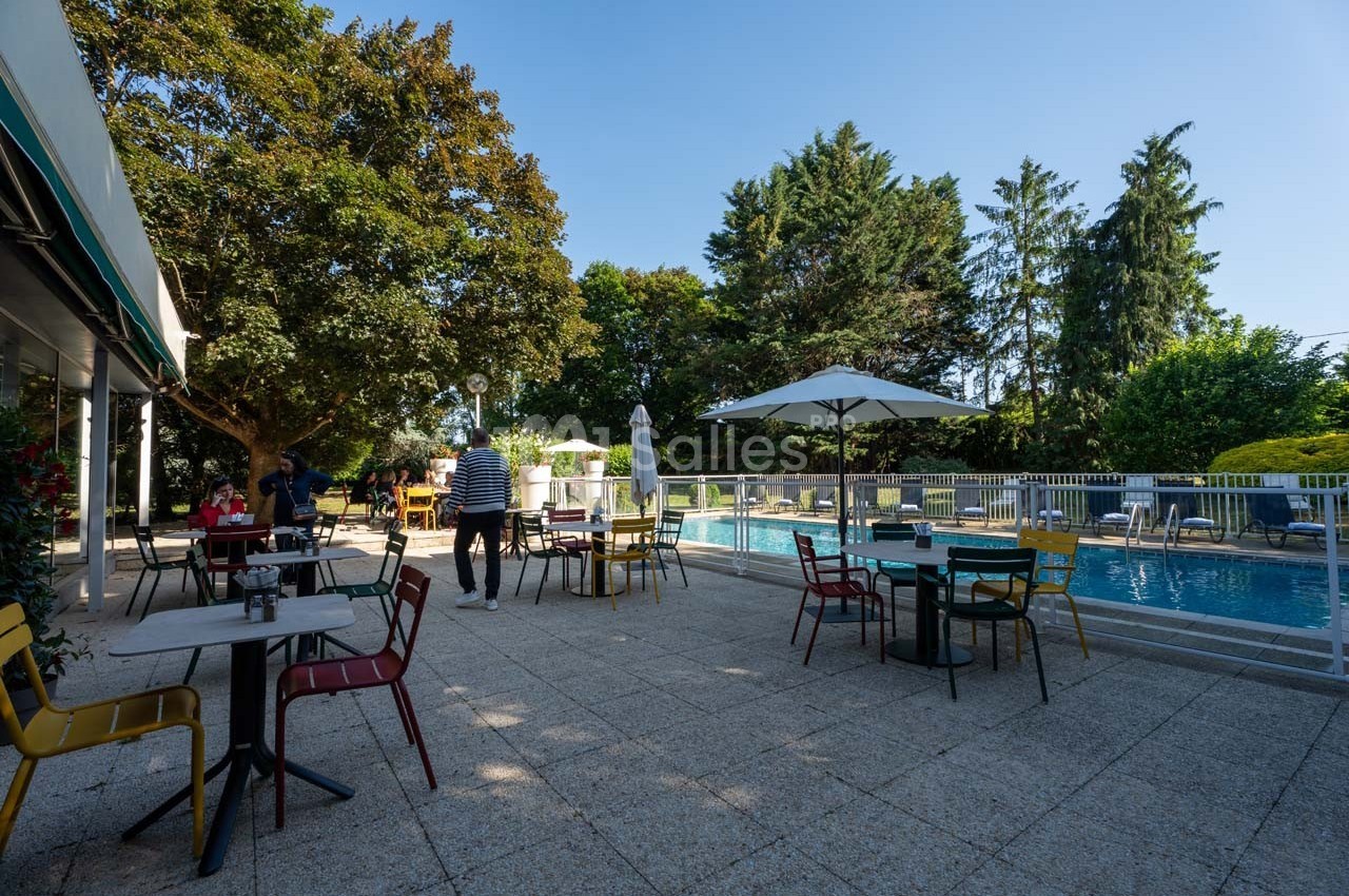 Terrasse avec tables et chaises colorées, bordée d'une piscine et entourée de végétation sous un ciel dégagé.