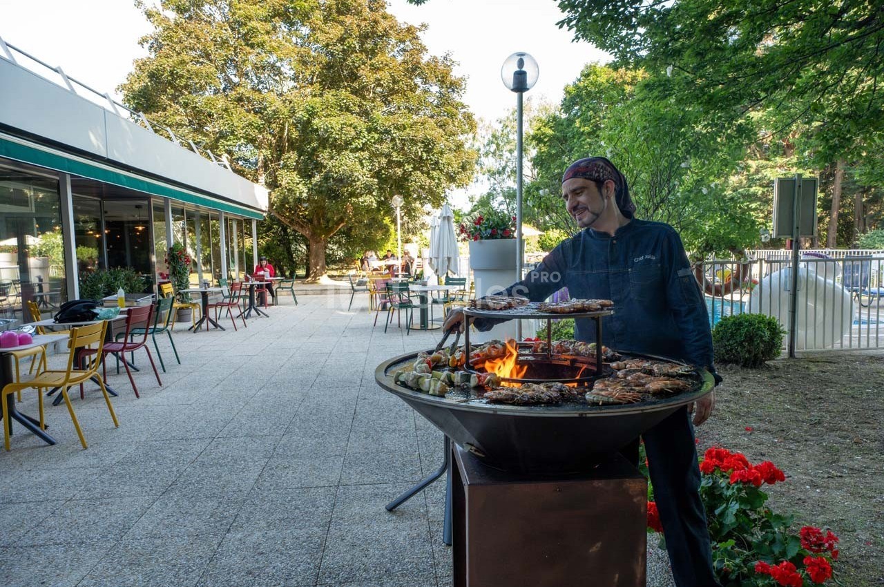 Un cuisinier prépare des grillades sur un barbecue en plein air près d'une terrasse de restaurant.