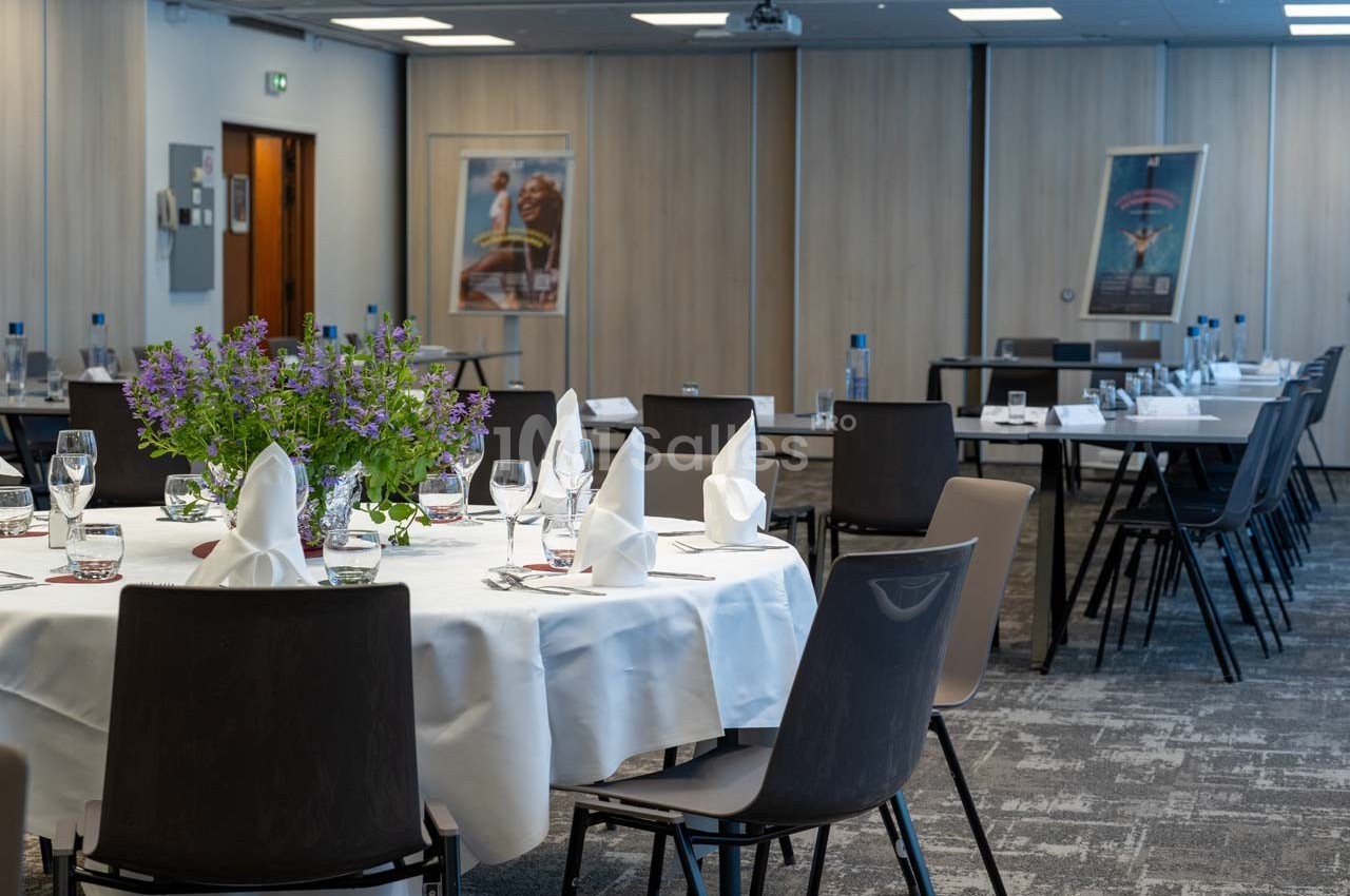 Salle de réception avec tables rondes dressées, nappes blanches, verres, serviettes pliées et décoration florale.