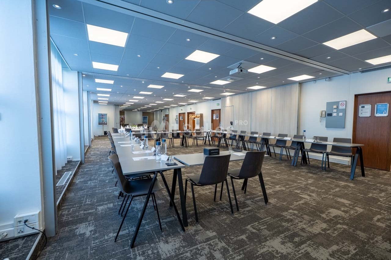 Salle de réunion lumineuse avec des tables disposées en U, des chaises noires et un sol en moquette grise.
