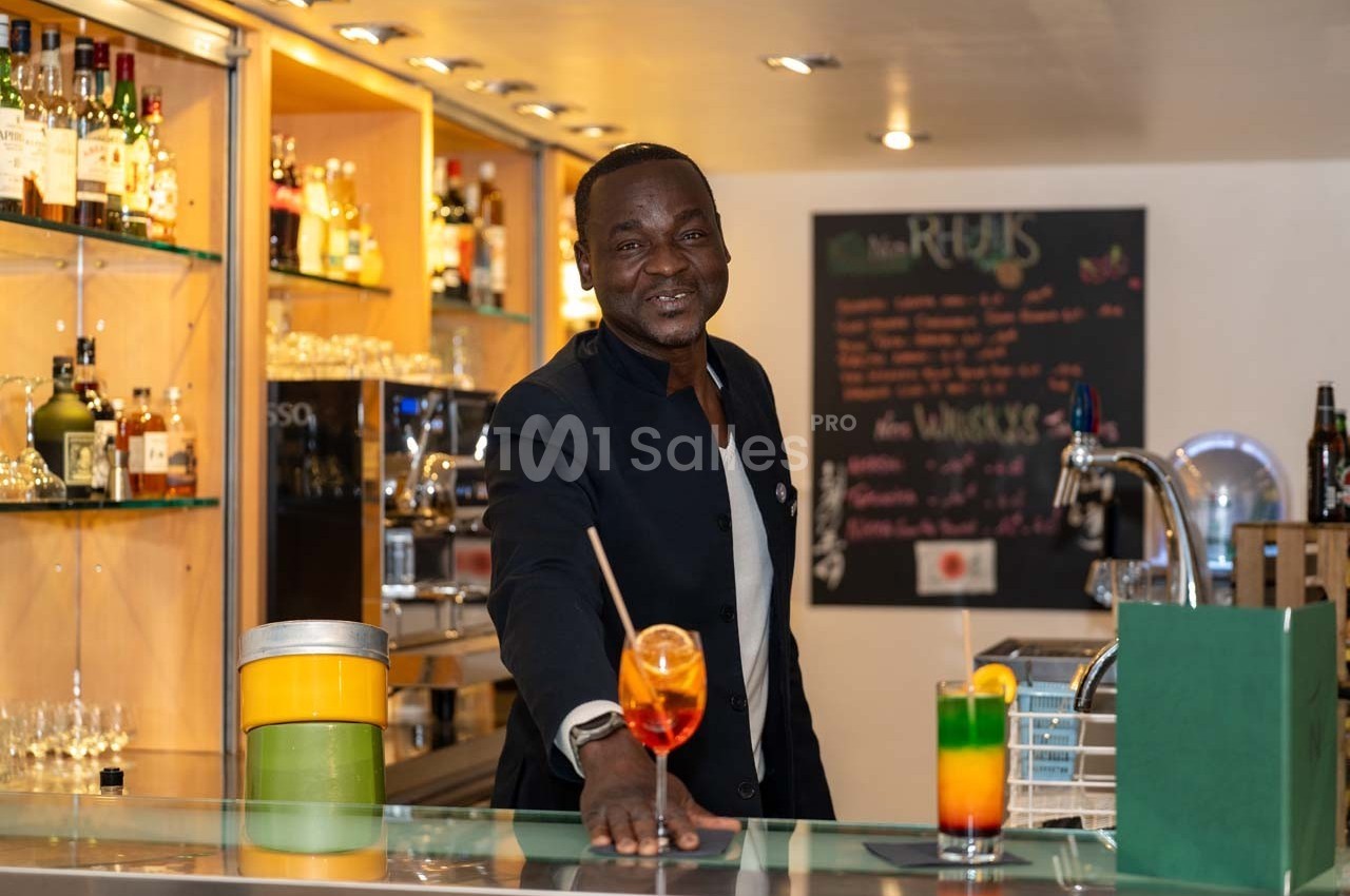 Un barman souriant présente un cocktail coloré sur le comptoir d'un bar bien éclairé avec des étagères de bouteilles.