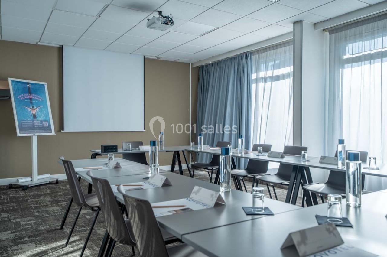 Salle de réunion lumineuse avec tables disposées en U, bouteilles d'eau, porte-noms et écran de projection.