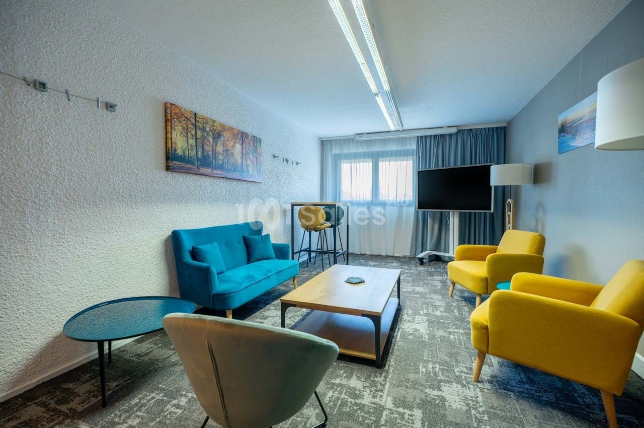 Salon moderne avec canapé bleu, fauteuils jaunes, table basse en bois, téléviseur et grande fenêtre avec rideaux.