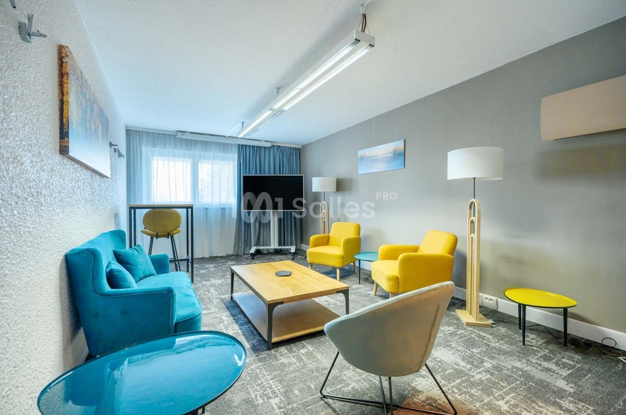 Salle de réunion lumineuse avec canapés bleus, fauteuils jaunes, table basse en bois et écran de télévision.