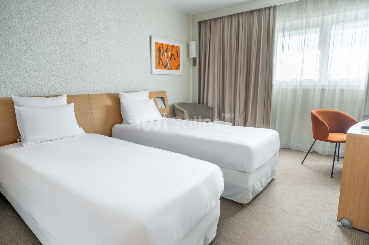 Chambre d'hôtel lumineuse avec deux lits simples, un fauteuil, un bureau et une grande fenêtre avec rideaux beiges.