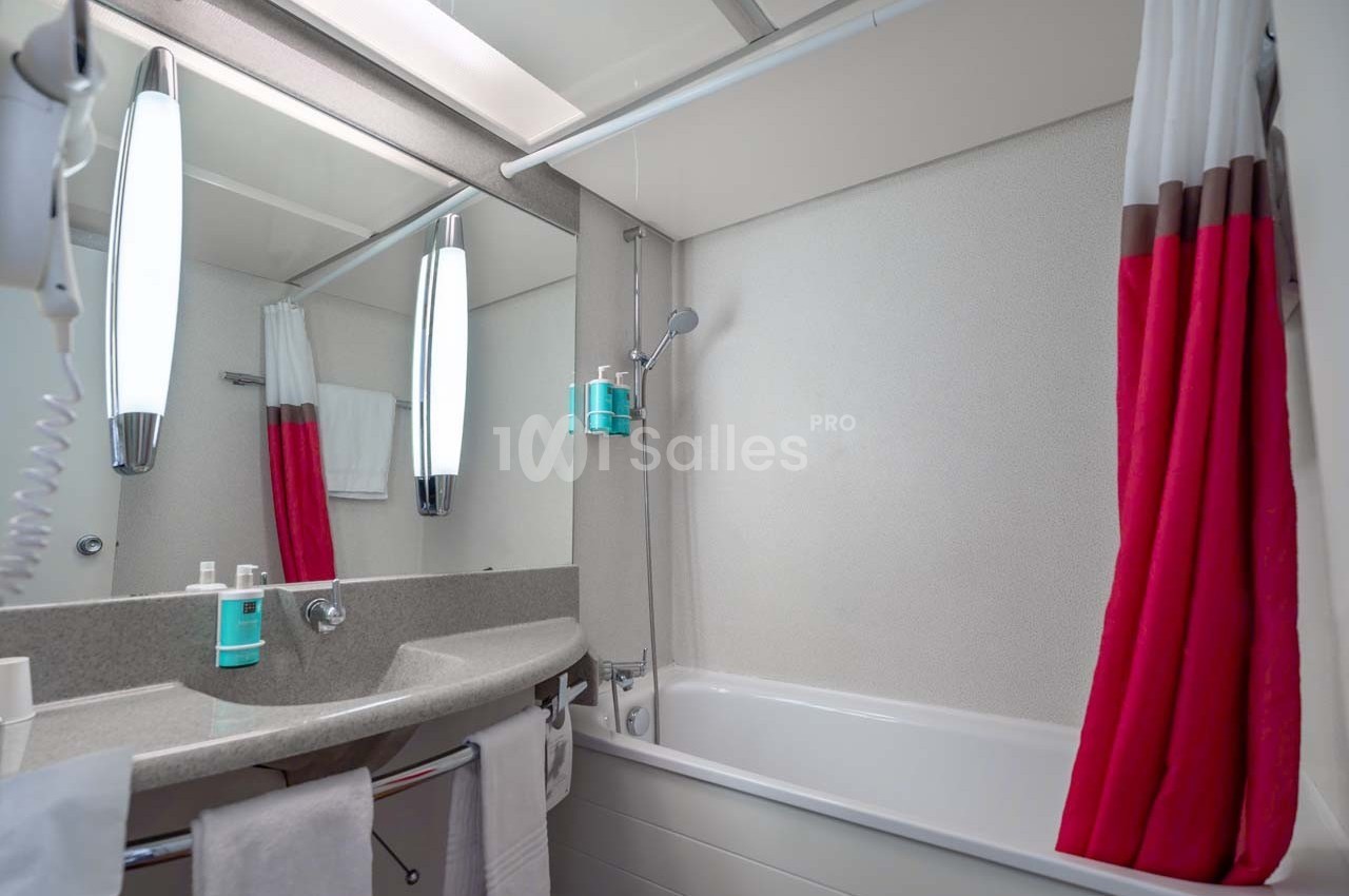 Salle de bain avec lavabo, miroir éclairé, baignoire équipée d'une douche et rideau rouge et blanc.