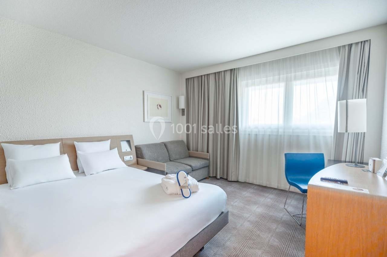 Chambre d'hôtel lumineuse avec lit double, canapé, bureau en bois, chaise bleue et grande fenêtre avec rideaux.