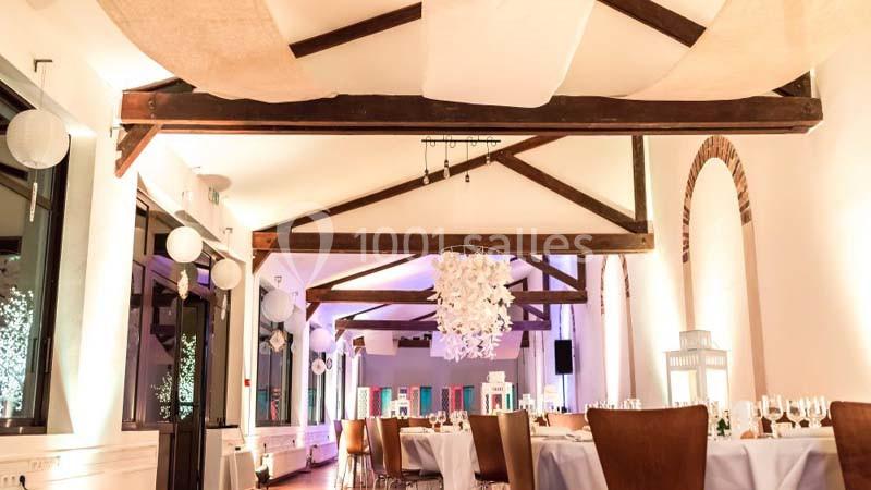 Salle de réception lumineuse avec poutres apparentes, tables dressées et décorations suspendues.