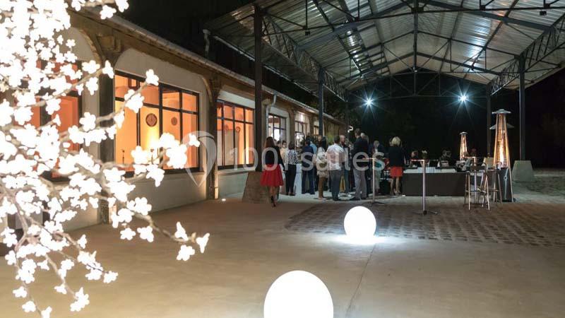 Groupe de personnes discutant sous une structure métallique éclairée, près d'un bâtiment avec de grandes fenêtres.