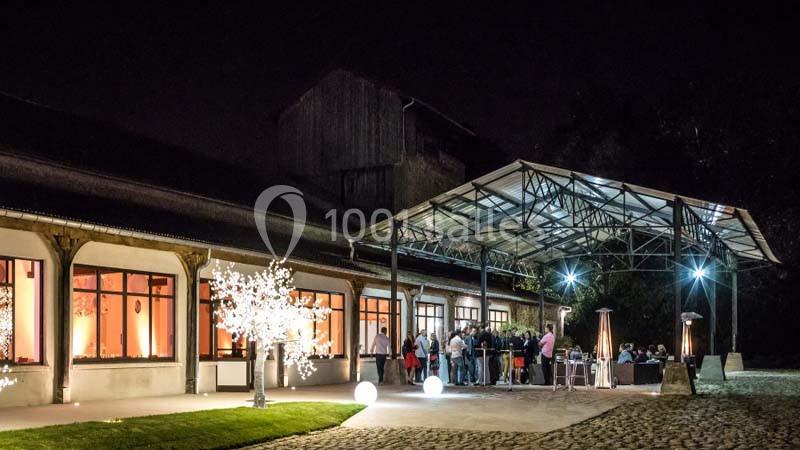 Vue nocturne d'un bâtiment éclairé avec une terrasse couverte, des invités rassemblés et un arbre lumineux décoratif.