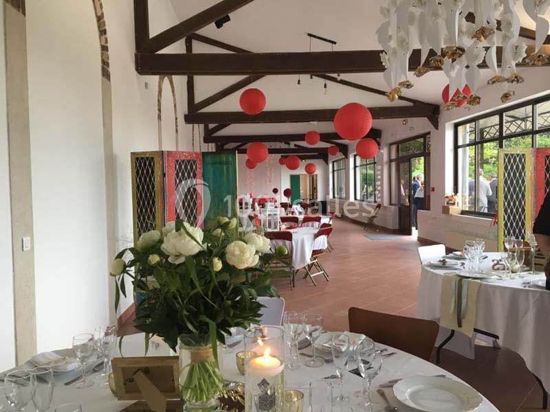 Salle de réception lumineuse décorée de lanternes rouges, tables dressées avec fleurs blanches et bougies.