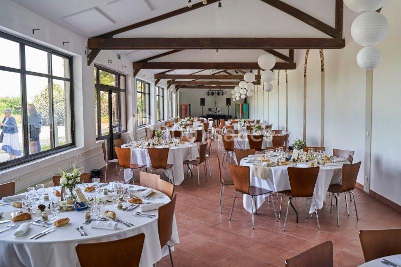 Salle de réception lumineuse avec tables rondes dressées, chaises en bois et décorations suspendues.