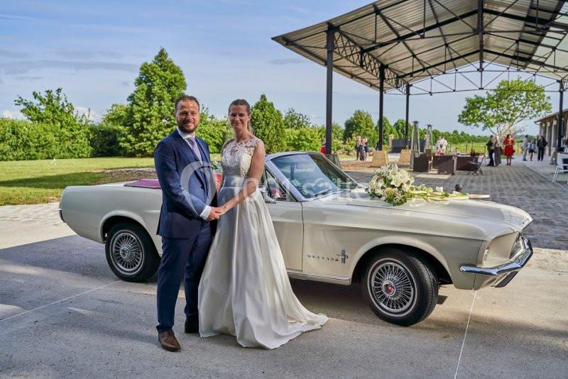 Un couple en tenue de mariage pose devant une voiture ancienne blanche, décorée de fleurs, dans un cadre extérieur.