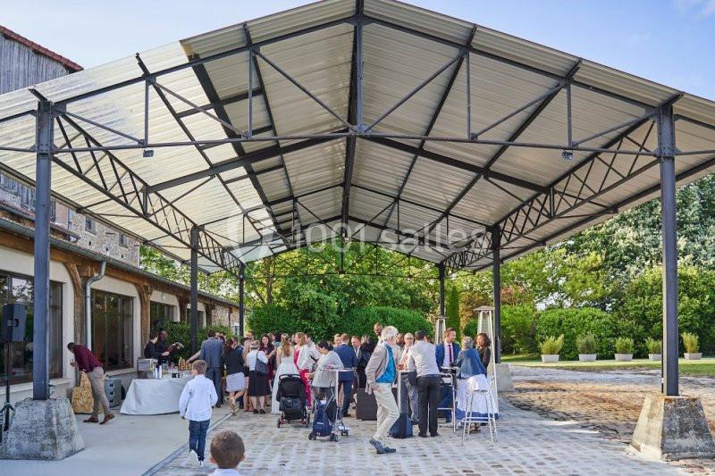 Groupe de personnes rassemblées sous une structure métallique en extérieur, près d'un bâtiment et d'espaces verts.